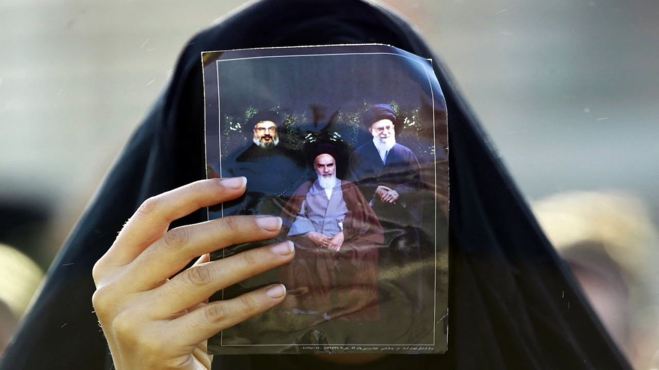 یک زائر مسلمان شیعه در جریان توقفی در نجف، در مسیر حرکت به‌سوی زیارتگاه شهر مرکزی کربلا، در ۹ دسامبر ۲۰۱۴، تصویری از علی خامنه‌ای (راست)، روح‌الله خمینی، بنیان‌گذار جمهوری اسلامی ایران (وسط)، و حسن نصرالله، رهبر جنبش شبه‌نظامی مسلمان شیعه حزب‌الله لبنان  را در دست دارد. عکس از حیدر حمدانی، خبرگزاری فرانسه.