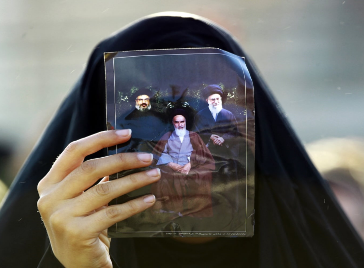 یک زائر مسلمان شیعه در جریان توقفی در نجف، در مسیر حرکت به‌سوی زیارتگاه شهر مرکزی کربلا، در ۹ دسامبر ۲۰۱۴، تصویری از علی خامنه‌ای (راست)، روح‌الله خمینی، بنیان‌گذار جمهوری اسلامی ایران (وسط)، و حسن نصرالله، رهبر جنبش شبه‌نظامی مسلمان شیعه حزب‌الله لبنان  را در دست دارد. عکس از حیدر حمدانی، خبرگزاری فرانسه.